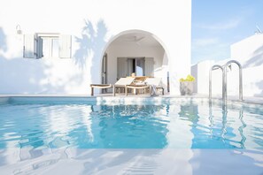 En klar och inbjudande pool med ett soligt uteområde, en vit byggnad med en välvd dörröppning och en metallstege som leder in i byggnaden.