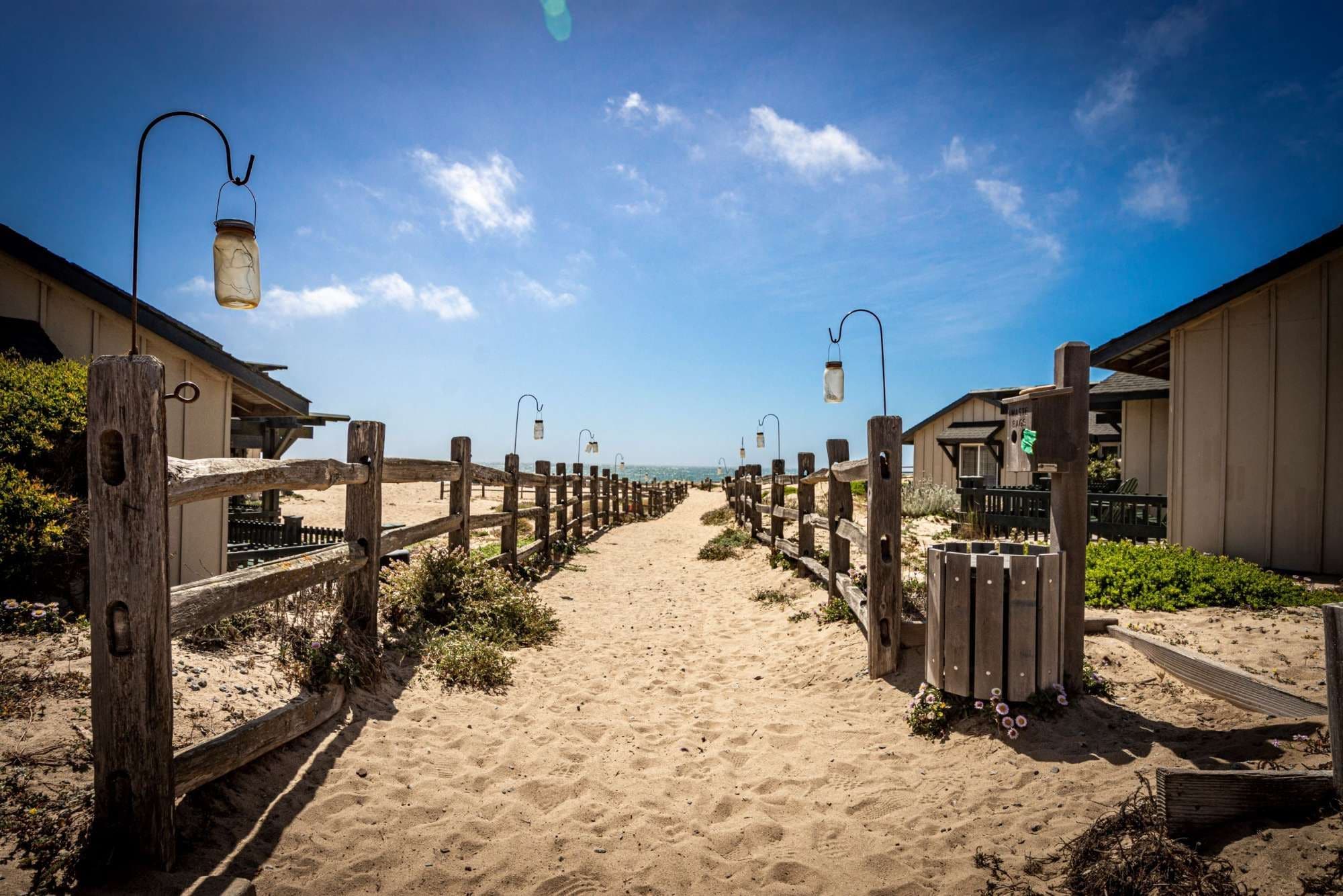 A sandy beach path flanked by wooden fences and beach huts with lanterns overhead.