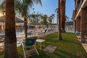 A pool area with lounge chairs and umbrellas, surrounded by palm trees and a building with a brick facade. - 3X3BR Desert Escape Near Coachella & amenities (Palm Springs)