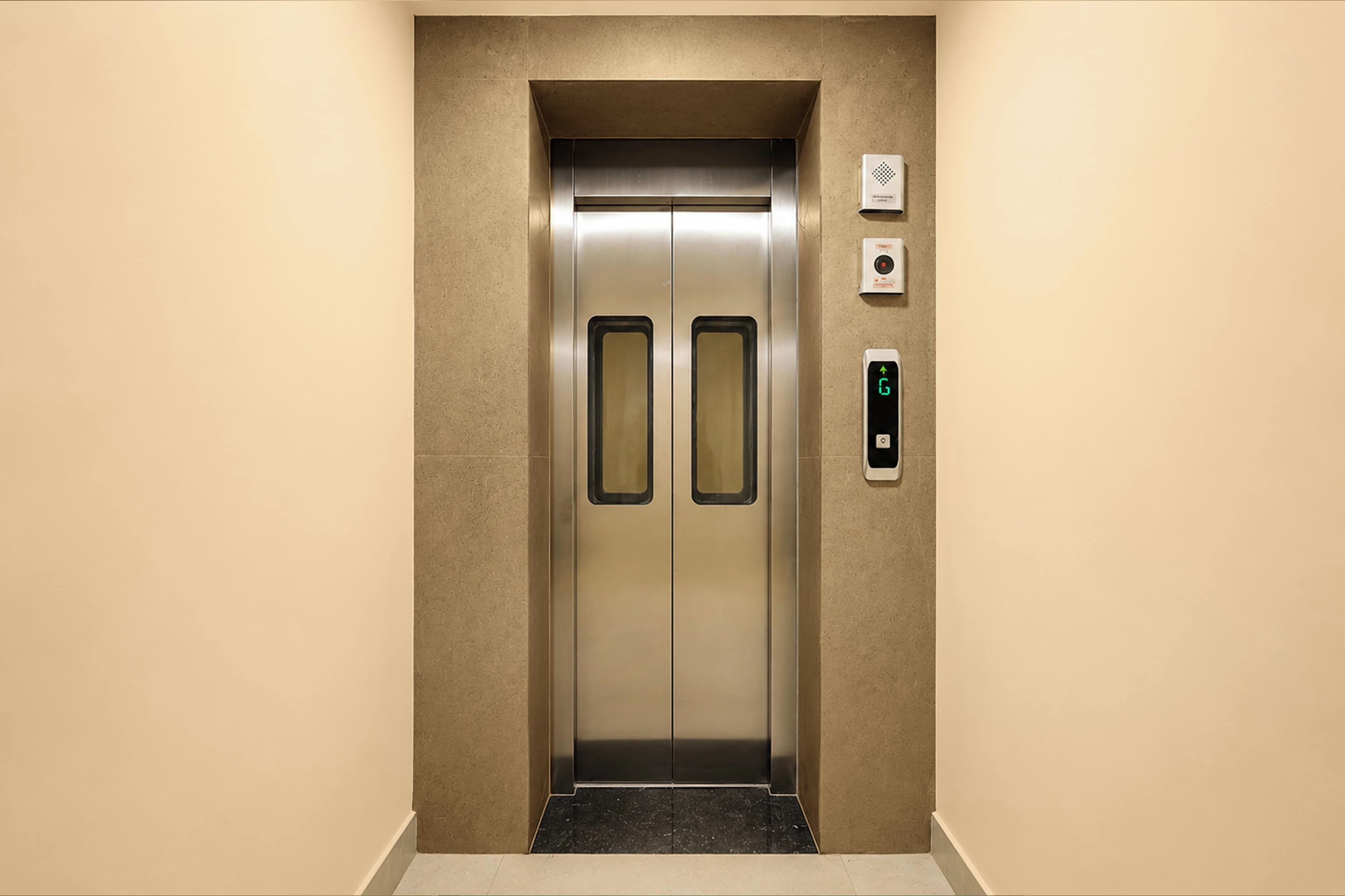 En elevator med metalfinish, et kontrolpanel med digital skærm og et intercomsystem monteret på væggen.