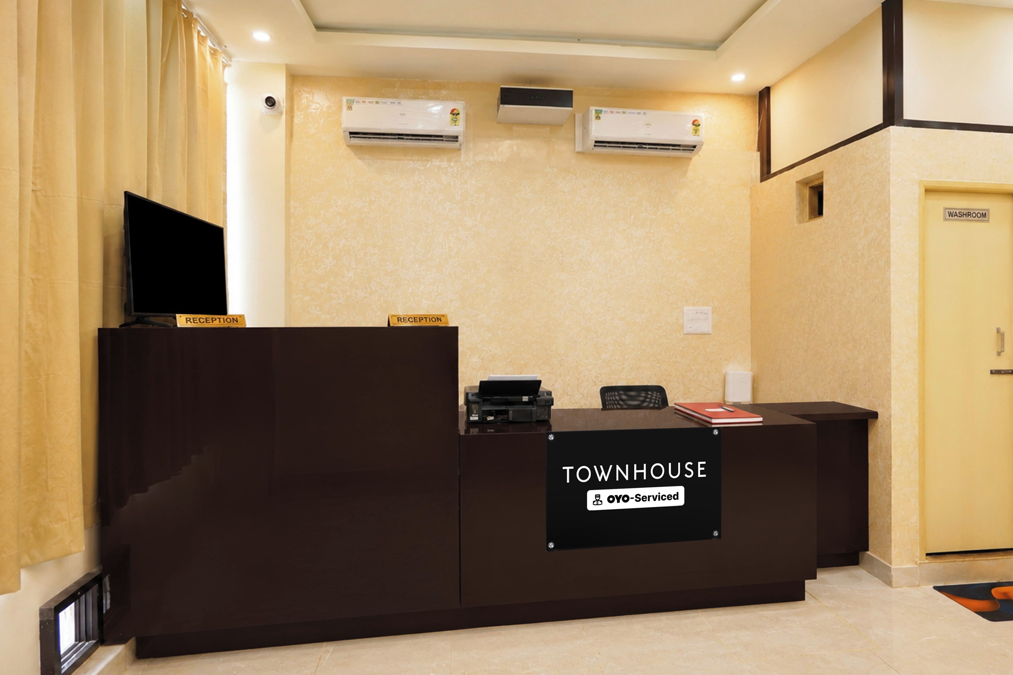 A reception desk with a 'TOWNHOUSE' sign, air conditioning units, a television, and a 'WASHROOM' door.