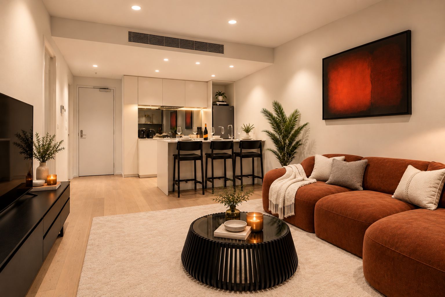 A modern living room with a sofa, a coffee table, and a TV. There is a kitchen area in the background with a dining table and chairs.