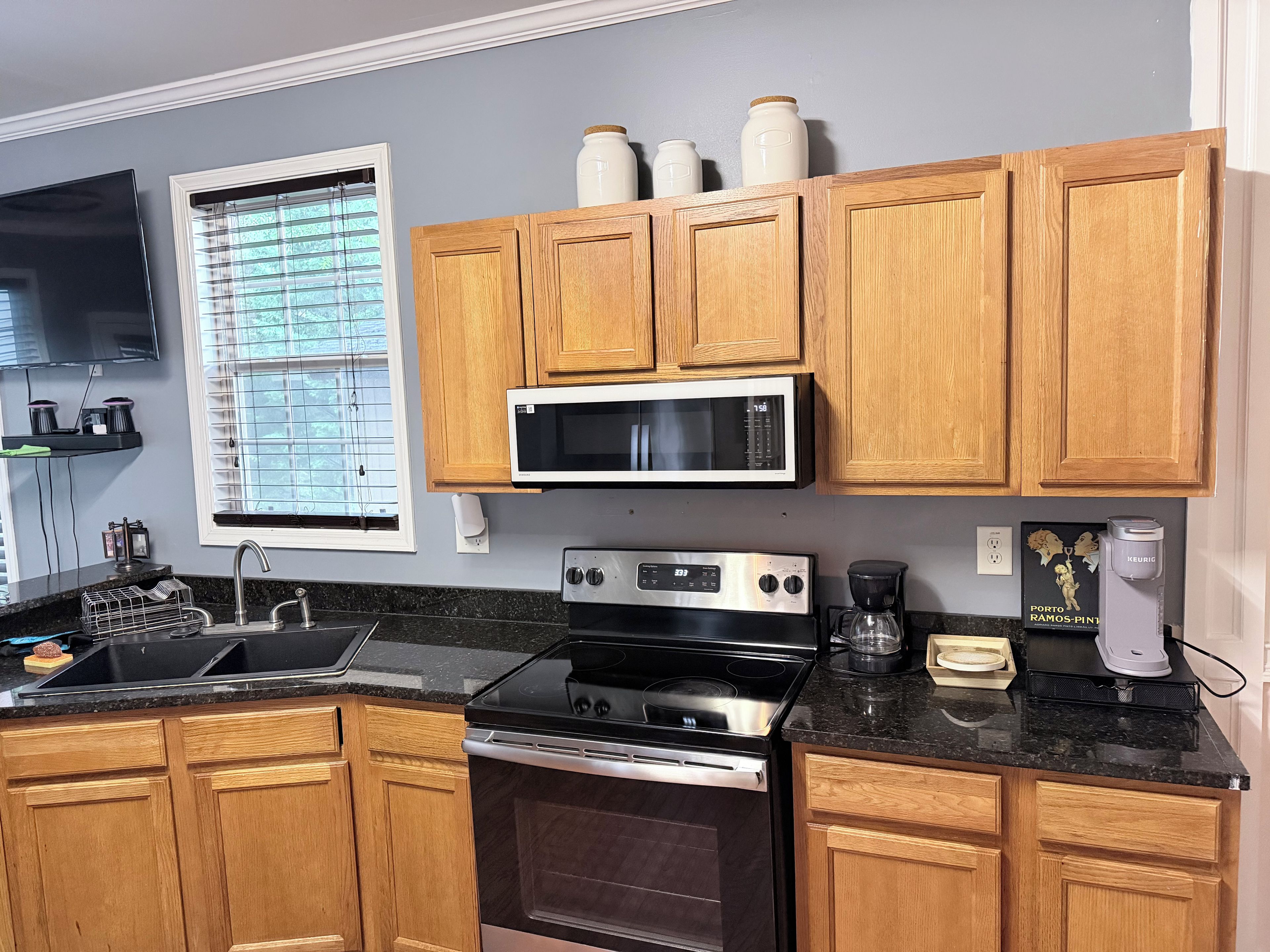 A kitchen with wooden cabinets, a microwave, an oven, a coffee maker, and a window with blinds.