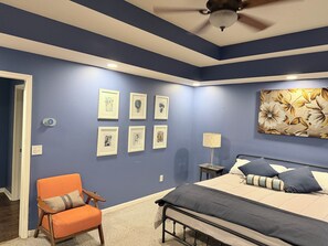 A bedroom with a bed, a chair, a ceiling fan, and wall-mounted artwork.