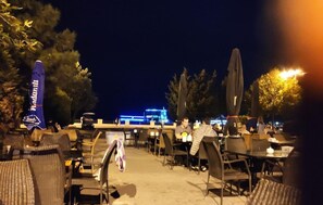 An outdoor dining area with tables and chairs, a flag, and a building with lights in the background. - Yacht Club (Baku)