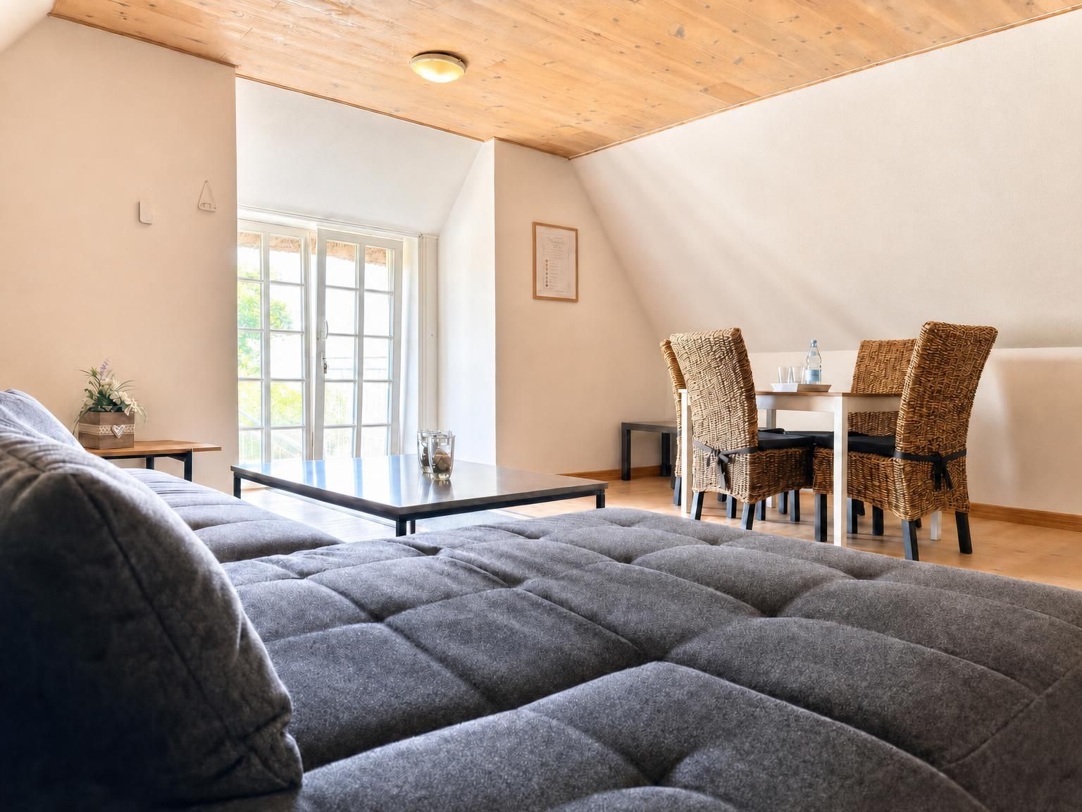 Un salon moderne avec un grand canapé gris, un coin repas avec des chaises en osier et un plafond en bois.