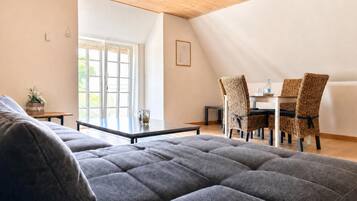 Un salon moderne avec un grand canapé gris, un coin repas avec des chaises en osier et un plafond en bois.