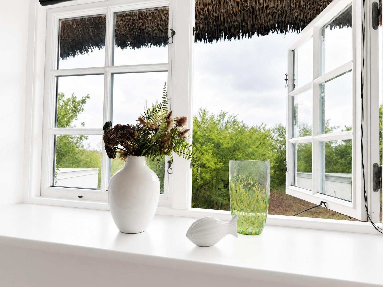 Eine weiße Fensterbank mit einer weißen Vase, die getrocknete Blumen enthält, sowie eine klare Glasvase mit grünem Grünzeug.