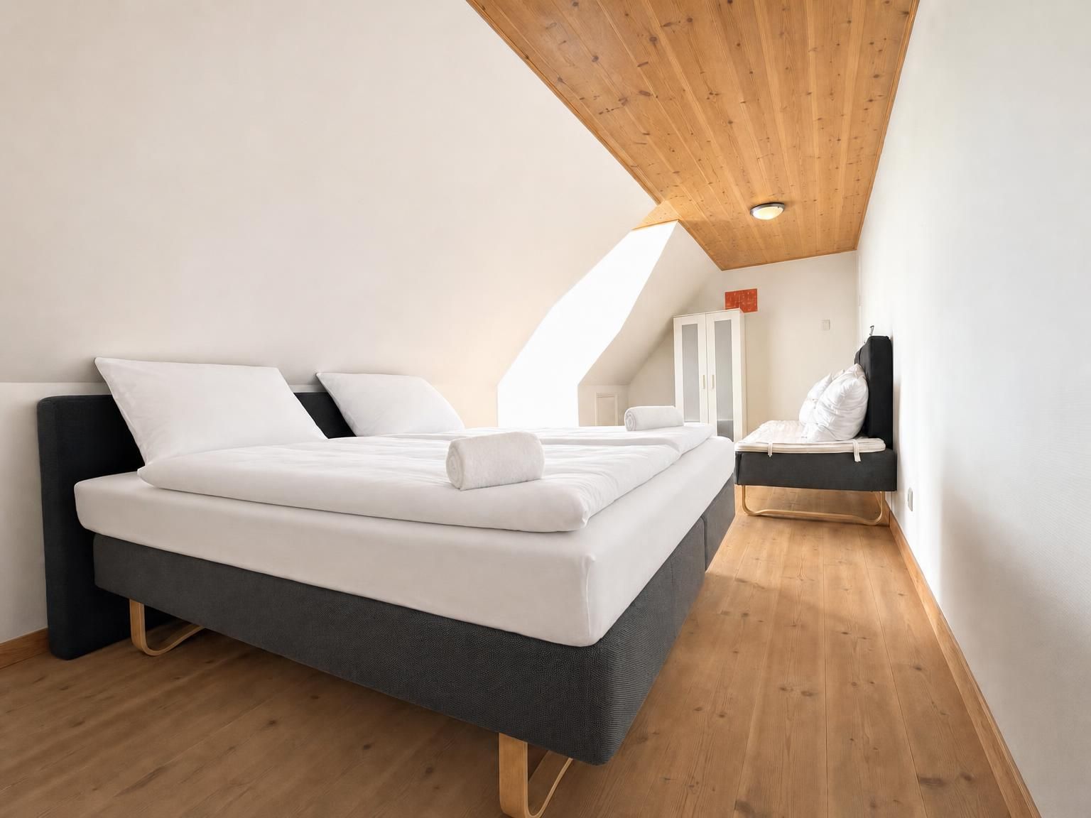 A modern bedroom with a large bed, a small seating area, and a sloped ceiling.