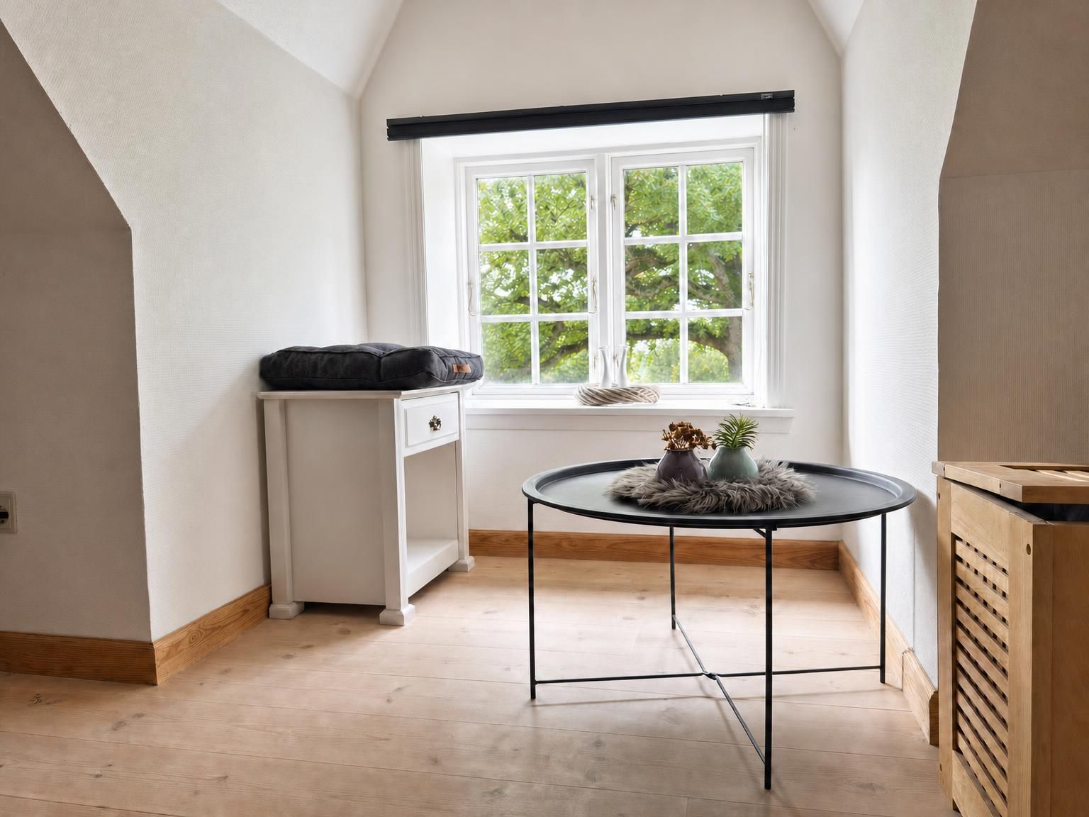 Una habitación con pared blanca, una ventana con vista a follaje, una pequeña mesa redonda con un florero y un cojín, y un armario blanco con un cojín negro encima.
