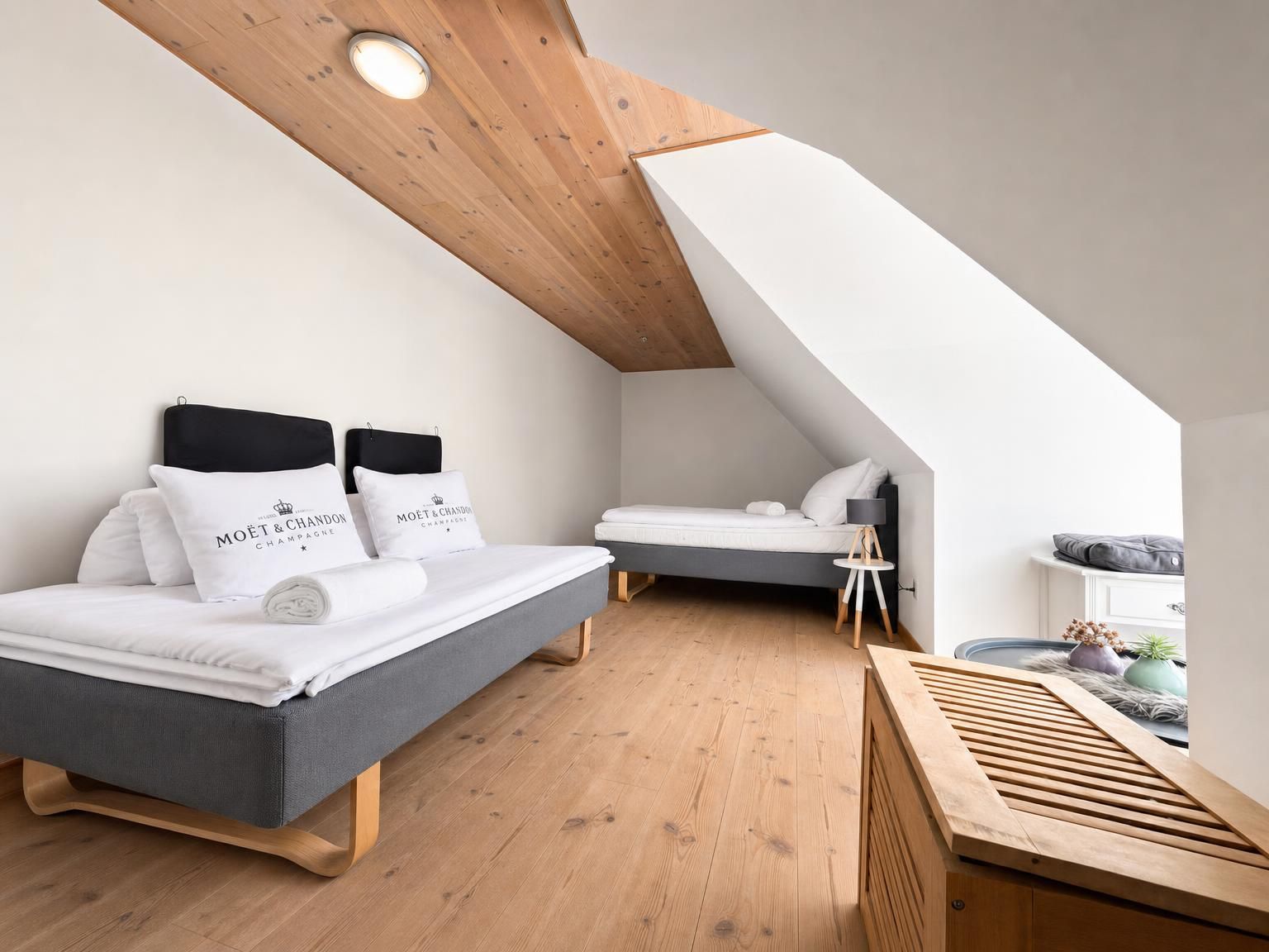 Ett modernt sovrum i loftstil med ett sluttande tak, en säng med vita sängkläder och kuddar med varumärkeslogotyp, ett litet sängbord och en träbänk.