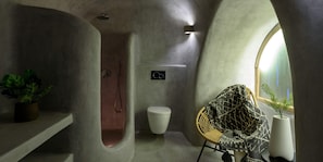 A bathroom with a curved shower, a toilet, and a chair with a patterned throw. - Sensation Villa By Senses Collection (Santorini)