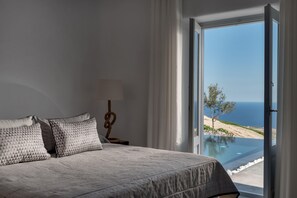 A bedroom with a bed, a nightstand, a lamp, and a view of the ocean through an open door. - Sensation Villa By Senses Collection (Santorini)