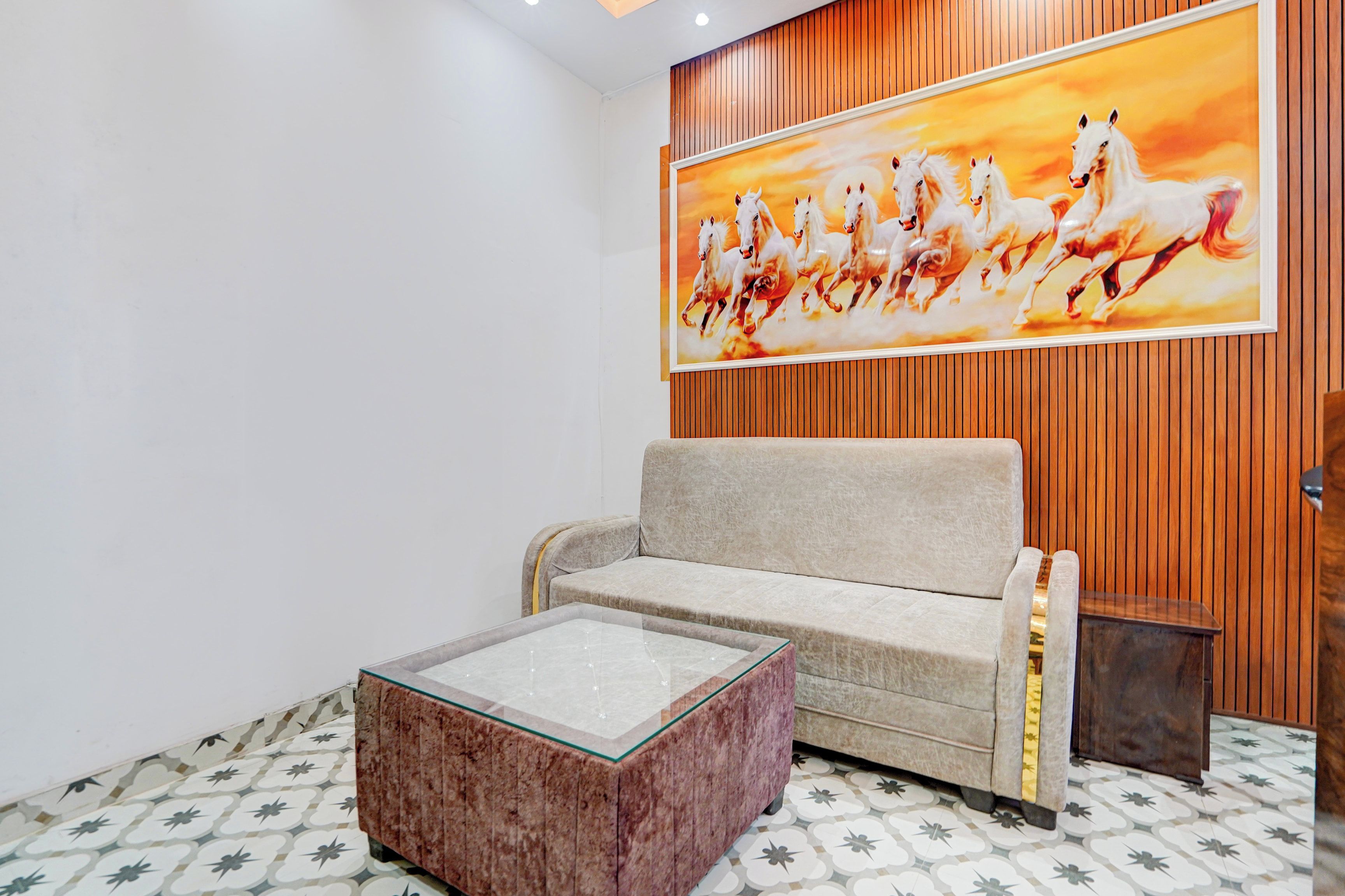 A room with a sofa, a glass coffee table, and a wooden panel wall with a painting of horses.