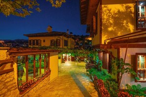 A courtyard with a pathway leading to a building entrance, surrounded by vegetation and a view of the surrounding area. - Cesmeli Konak (Safranbolu)