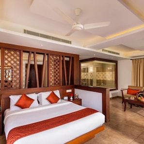 Habitación de hotel con cama, escritorio, silla, ventilador de techo y baño visible a través de una mampara de vidrio.