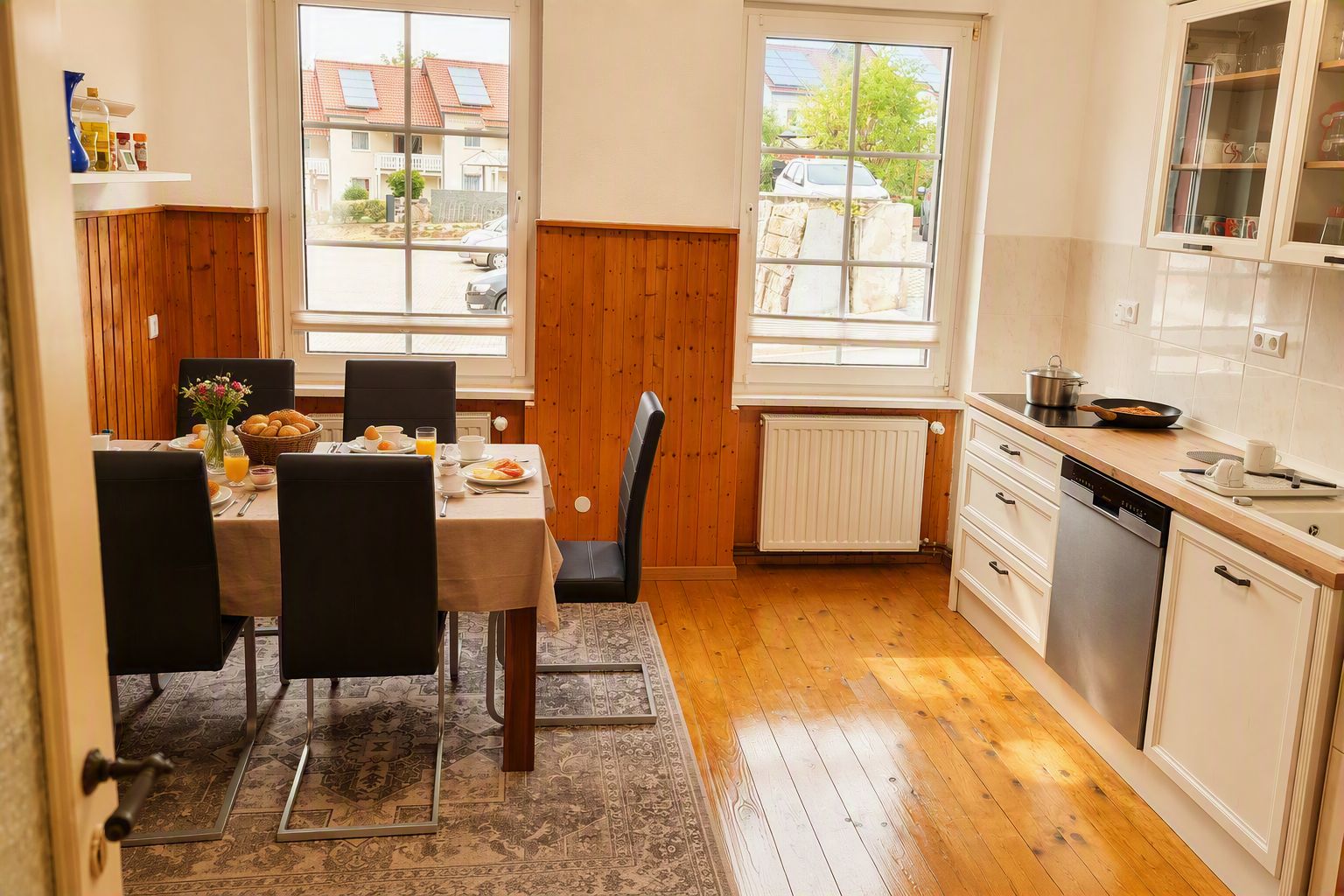Ein Essbereich mit einem Tisch für zwei Personen, eine Küche mit weißen Schränken und einem Geschirrspüler sowie große Fenster mit Blick nach draußen.