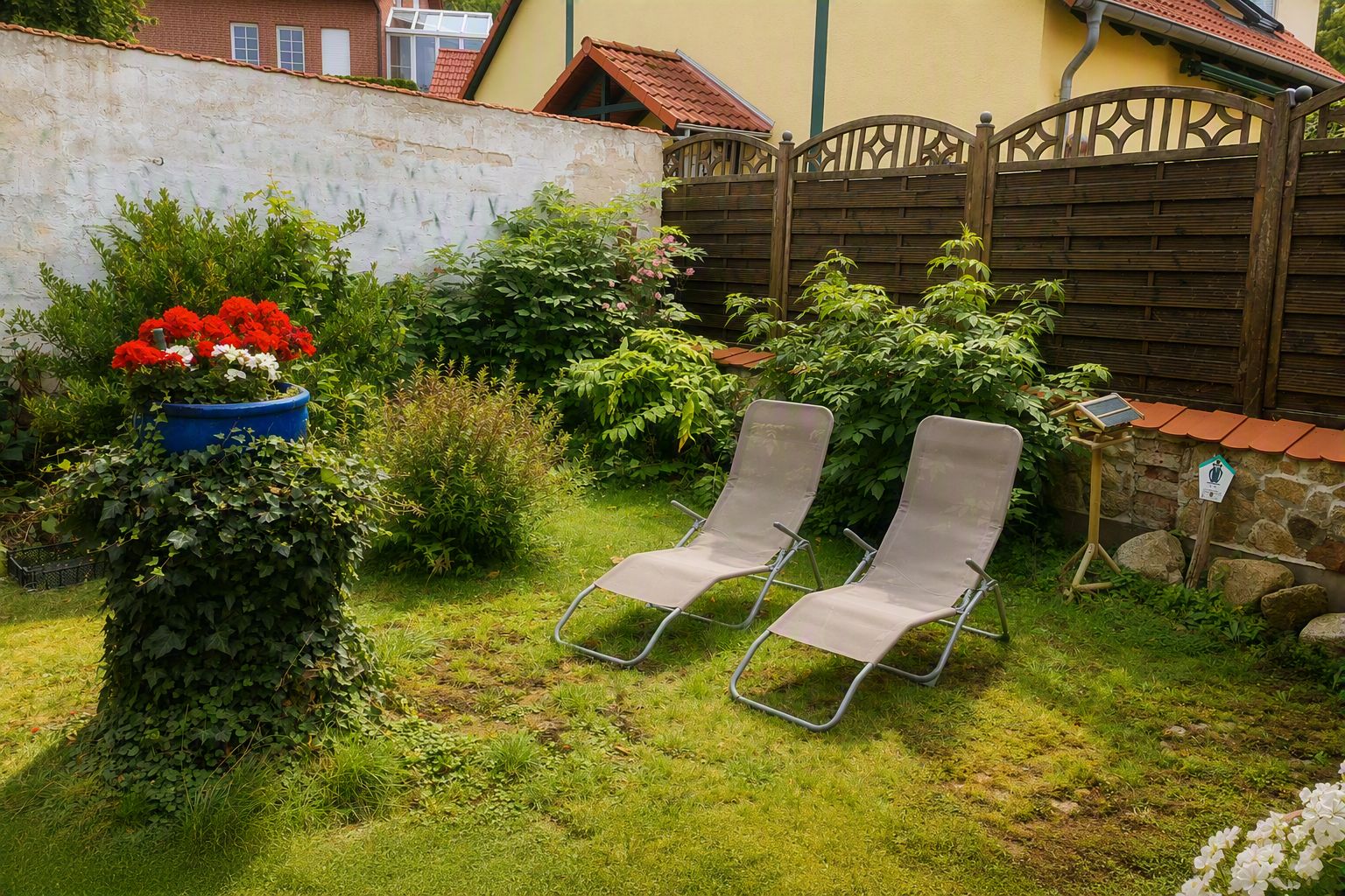 Ein Garten mit zwei Metallstühlen, einem blauen Blumenkasten mit roten Blüten und einem Holzzaun.