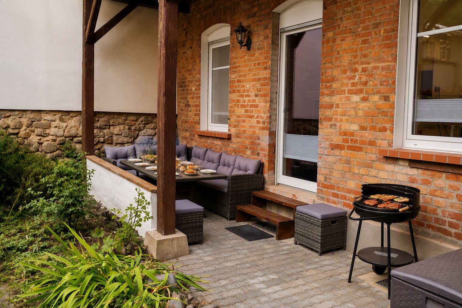 An outdoor patio with a brick wall, a table set for dining, a barbecue grill, and a seating area with cushions.