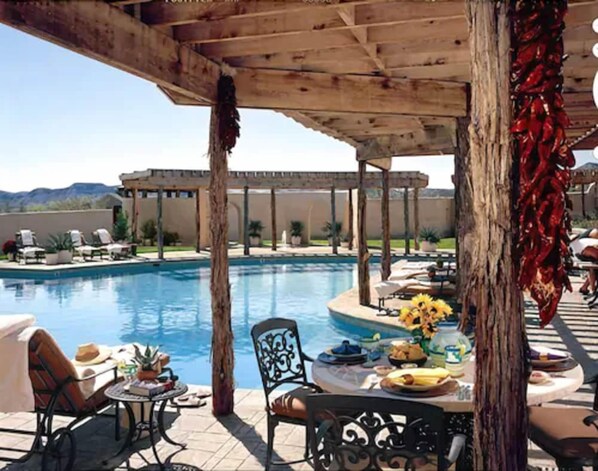 Zona de piscina con área de estar cubierta, una mesa con comida y bebidas, y vistas a la piscina y el paisaje.