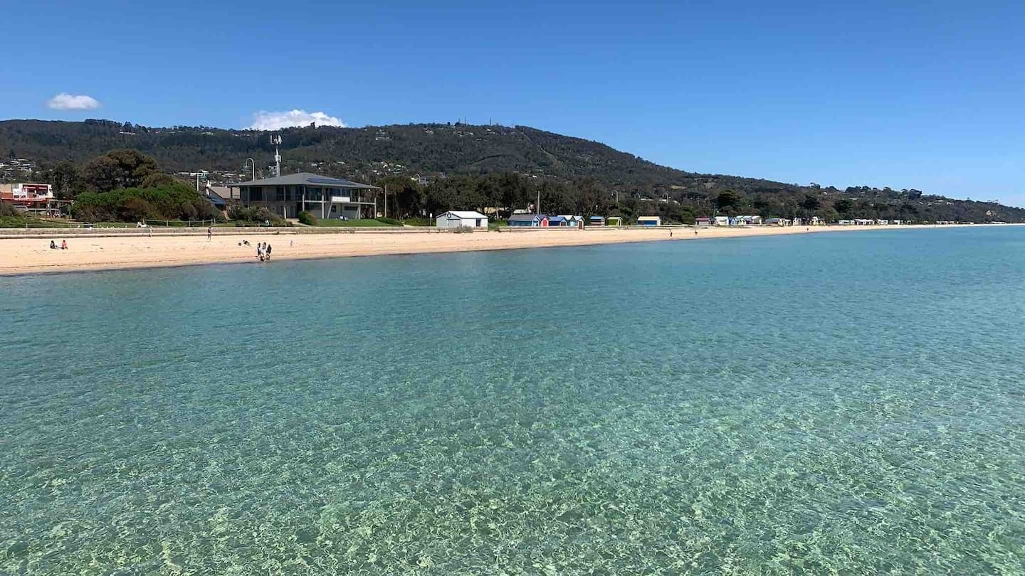 Una playa costera con agua cristalina, un edificio de techo plano y un paisaje montañoso al fondo.