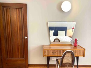 Ein Zimmer mit einer Holztür, einem Bett mit blauer Kopfteil, einem Nachttisch mit einer roten Vase und einem Sessel.