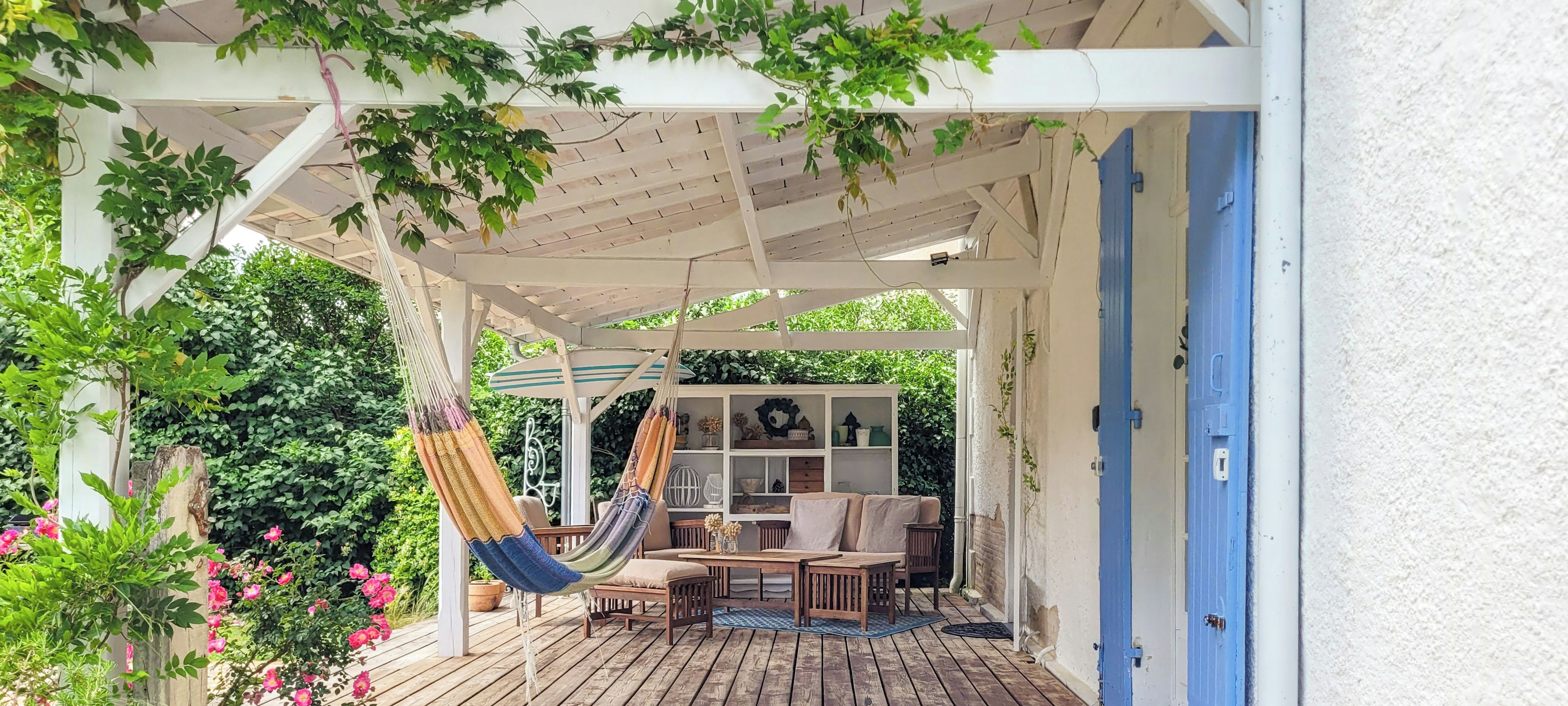 A wooden deck with a hammock, a sofa, and a small table, surrounded by greenery and a white pergola.
