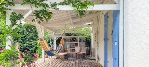 Una terraza de madera con una hamaca, un sofá y una mesita, rodeada de follaje y una pérgola blanca.