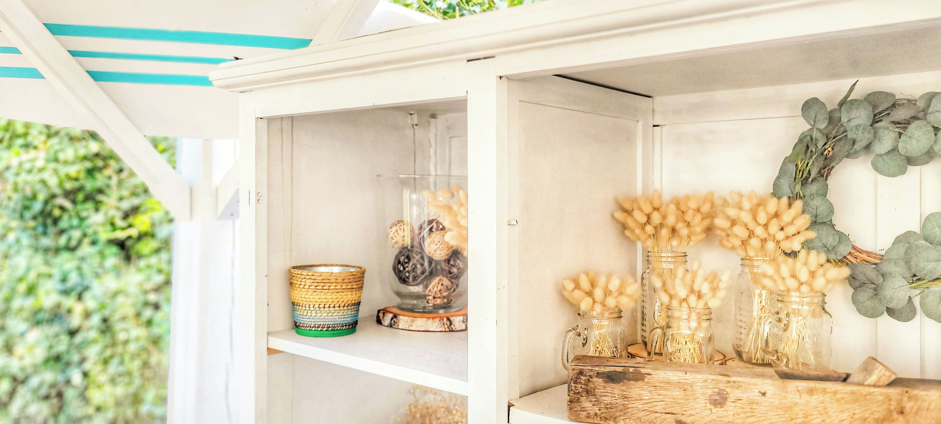Un estante blanco con objetos decorativos, incluyendo una cesta tejida, un florero de vidrio con un adorno y varios frascos de vidrio con material esponjoso.