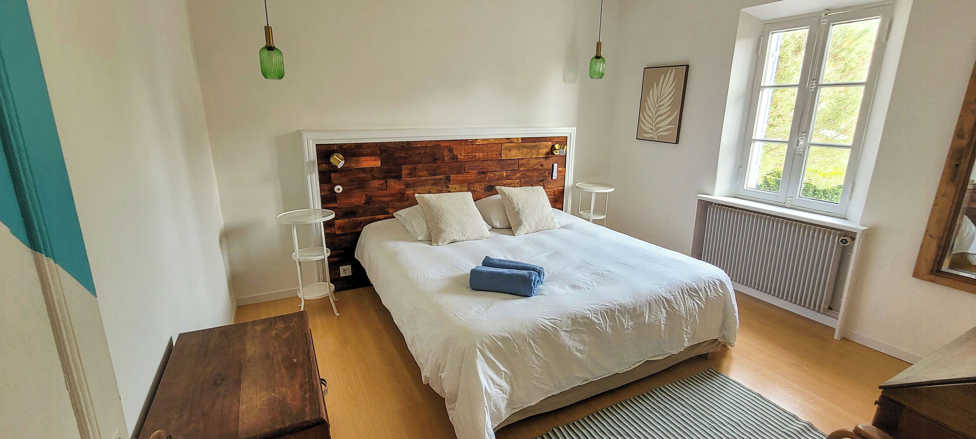 Une chambre à coucher avec une tête de lit en bois, un lit avec une literie blanche, deux lampes de chevet, une fenêtre donnant sur un paysage agréable et un radiateur.