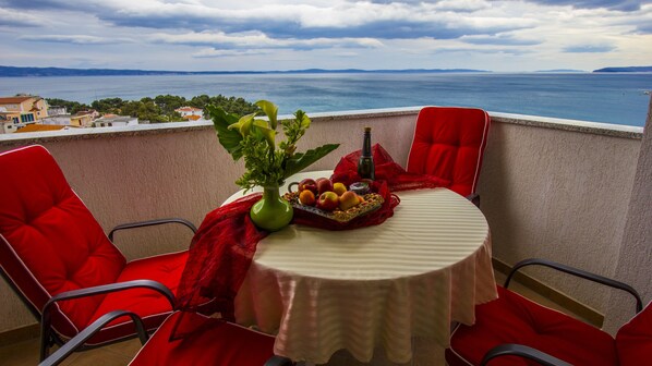 Een balkon met een tafel gedekt voor twee personen, met een bloevas, een fles en een fruitschaal. Er is uitzicht op een waterpartij en het landschap in de verte.