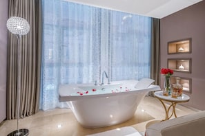 A modern bathroom with a freestanding bathtub, a large window with sheer curtains, a gold-framed side table with decorative items, and a chandelier. - Crystal Orange Hotel (Tianjin)