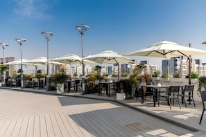 Outdoor seating with umbrellas, tables, and chairs, set against a cityscape backdrop.
