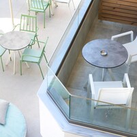 A balcony with a glass railing, a round table with a metal bowl, and green and white chairs.