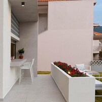 Un balcón moderno al aire libre con baranda blanca, zona de estar con sillas blancas y maceta con flores rojas.