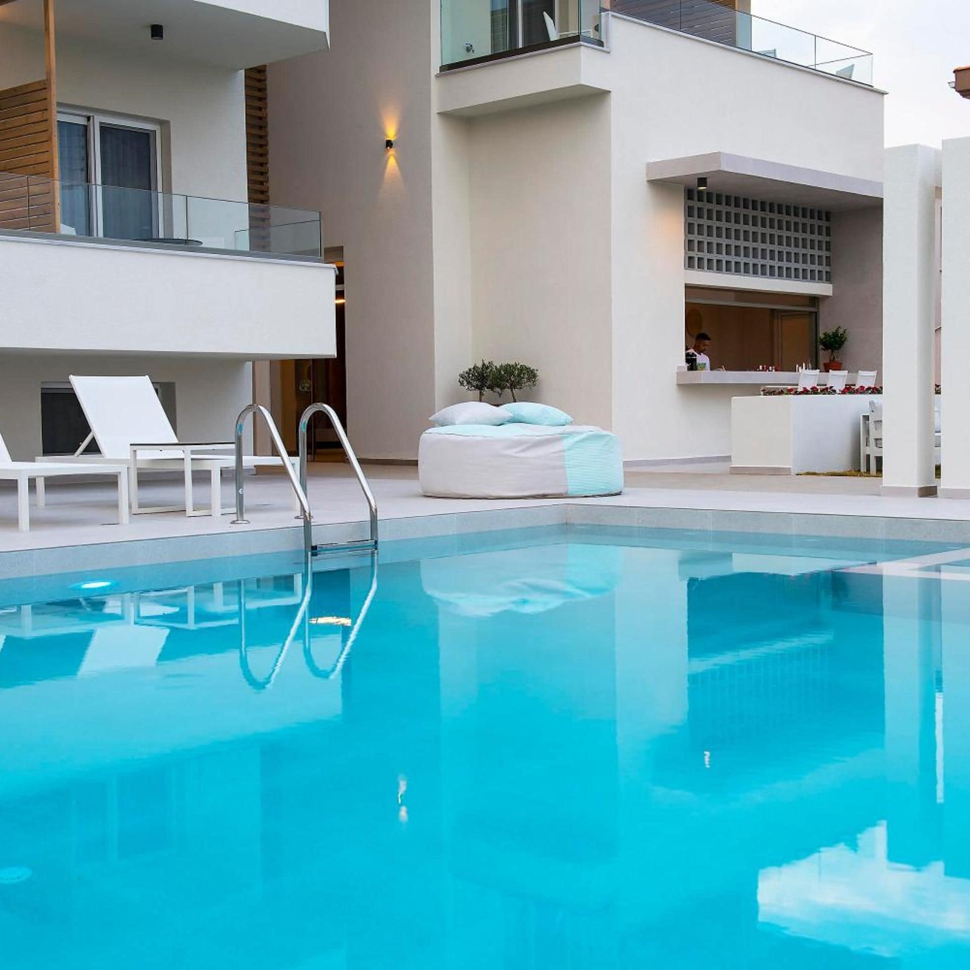 Zona de piscina moderna de hotel con piscina cristalina, sillas de descanso y un mostrador de recepción.