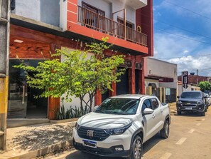 Un SUV blanco FIAT estacionado en una calle con un edificio que tiene un balcón y un cartel que dice 'VELAS'.