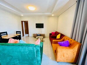 A living room with a sofa, a dining table, and a TV mounted on the wall. - Convenient 2BR apartment near kigali airport (Kigali)
