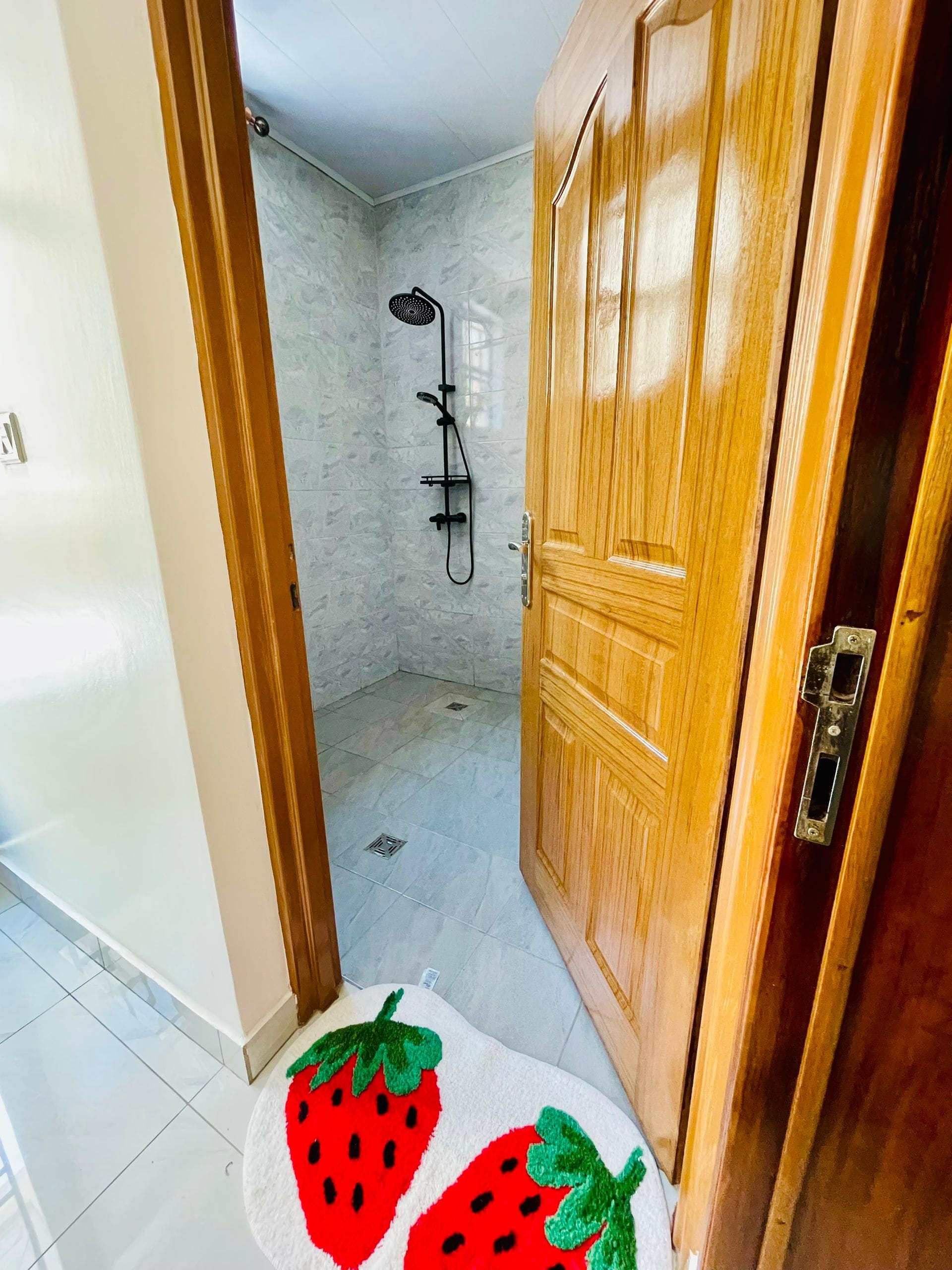 Un corridoio con una porta in legno, uno spazio doccia con una ventola nera e un tappeto a tema fragola.