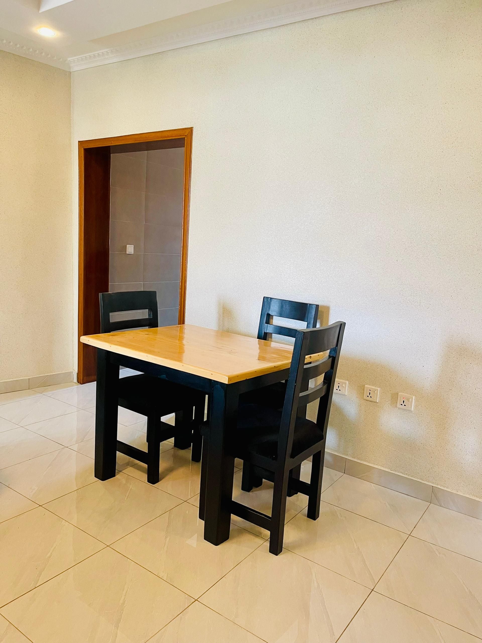 A dining area with a wooden table and four chairs, a tiled floor, and a plain wall with electrical outlets.