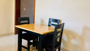 A dining area with a wooden table and four chairs, a tiled floor, and a plain wall with electrical outlets.