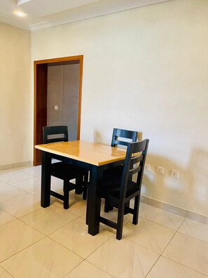A dining area with a wooden table and four chairs, a tiled floor, and a plain wall with electrical outlets. - Convenient 2BR apartment near kigali airport (Kigali)
