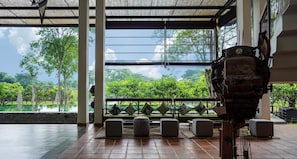A room with a large window overlooking a lush garden and a pool. There are several cushioned seating areas and a large, ornate mechanical object in the foreground. - Amba Yaalu Kandalama (Kandalama)