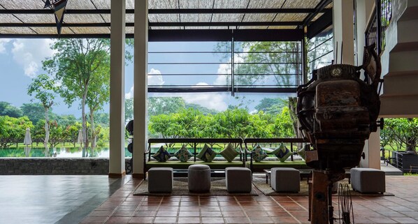 Habitació amb una finestra gran que dona a un jardí exuberant i una piscina. Hi ha diverses zones d'estar amb seients mullidors i un objecte mecànic gran i ornamentat en primer pla.