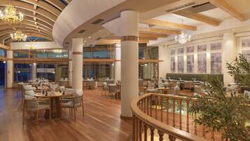 A spacious hotel dining area with multiple tables and chairs, large windows, and a wooden staircase.