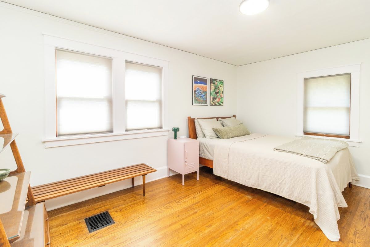 Un dormitorio con cama, una banca, una mesita de noche y dos ventanas.