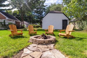 Un patio trasero con una fogata, cuatro sillas Adirondack y una pequeña construcción blanca.