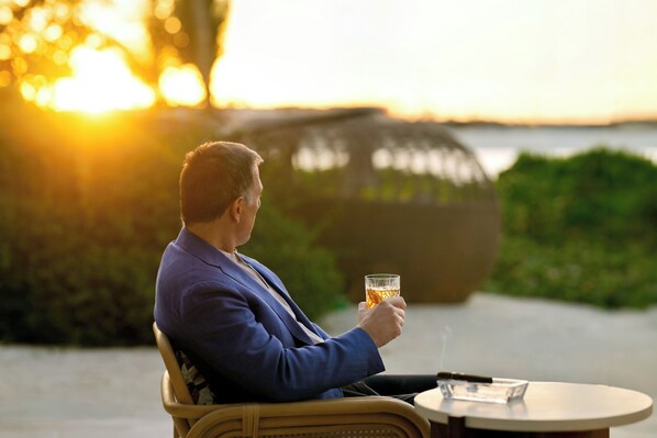 A person seated at a table with a drink, overlooking a body of water and greenery.