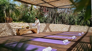 Een houten terras met yogamatten, een tafel met drinken en fruit, en een persoon in het wit.