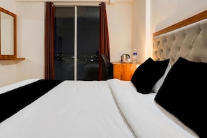 A hotel room with a bed, a nightstand, a chair, a window with curtains, and a view of the city at night. - Super Townhouse Lohegaon Near Pune Airport Formerly Landmark Regency (Pune)